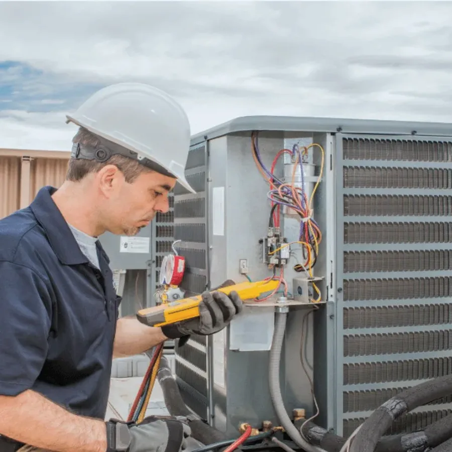 HVAC technician in a hard hat testing an outdoor air conditioner with a multimeter